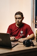 A person wearing a red shirt is sitting at a table, using a stylus on a digital tablet. A laptop is open in front of them, and a camera is placed on the table nearby. The setting appears to be indoors with soft lighting.