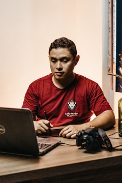 A person wearing a red shirt is sitting at a table, using a stylus on a digital tablet. A laptop is open in front of them, and a camera is placed on the table nearby. The setting appears to be indoors with soft lighting.