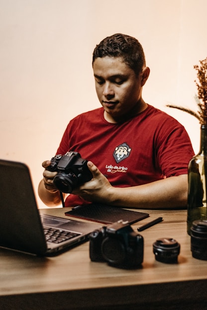A person wearing a red shirt sits at a wooden desk, holding a DSLR camera. On the desk are a laptop, a tablet with a stylus, an additional camera lens, and a decorative plant in a bottle. The lighting is warm and creates a cozy atmosphere.