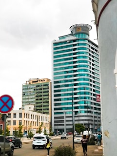 A modern cityscape with a prominent glass skyscraper featuring turquoise windows, surrounded by several other buildings. People walk along the street, and there are multiple cars on the road. The weather appears overcast, with a cloudy sky.