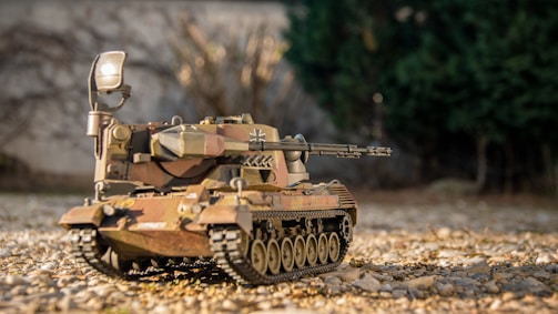 A detailed model of a military tank is shown, placed on a surface covered with small pebbles. The tank features a variety of intricate details including camouflage paint, realistic treads, and a mounted gun. The background is slightly blurred, suggesting a natural or outdoor setting with trees and foliage.