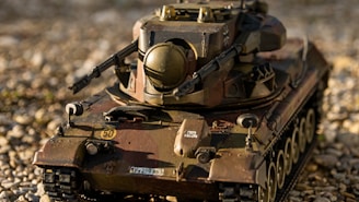 A detailed model of a military tank is displayed on a gravel surface. The tank features realistic details, including a turret, gun barrel, and tracks. The model has a weathered appearance with camouflaged paint in shades of green, brown, and black. Small details like numbers and insignias are visible on the body.
