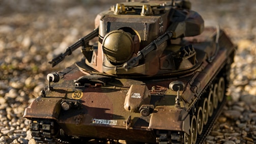 A detailed model of a military tank is displayed on a gravel surface. The tank features realistic details, including a turret, gun barrel, and tracks. The model has a weathered appearance with camouflaged paint in shades of green, brown, and black. Small details like numbers and insignias are visible on the body.