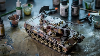 A detailed model tank is placed on a workbench surrounded by various paint bottles and tools. The tank has intricate details and a camouflage pattern. The surface has scattered paint marks indicating recent work.
