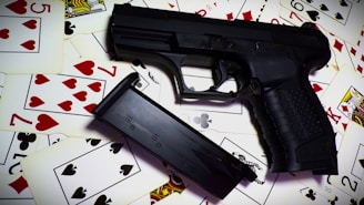 A black handgun and its magazine are placed on top of a scattered pile of playing cards. The cards display various suits and numbers, predominantly in red and black colors.