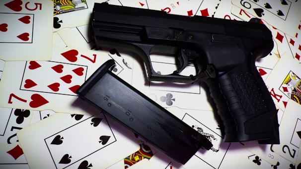 A black handgun and its magazine are placed on top of a scattered pile of playing cards. The cards display various suits and numbers, predominantly in red and black colors.