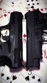 Two black firearms are placed on top of scattered playing cards. The cards include various suits and numbers, creating a contrasting backdrop for the guns.