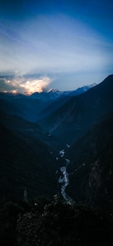A serene mountain landscape with a river winding through a deep valley under a sky transitioning from blue to golden hues at sunset. Snow-capped peaks are visible in the distance.
