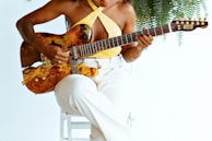 A person wearing a sleeveless yellow top and white pants plays a guitar with a unique, artistic design on its body while sitting on a white chair. Fern leaves hang in the background, complementing the artistic and serene setting.