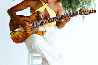 A person wearing a sleeveless yellow top and white pants plays a guitar with a unique, artistic design on its body while sitting on a white chair. Fern leaves hang in the background, complementing the artistic and serene setting.
