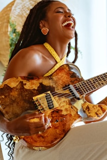 A person with long hair and heart-shaped earrings is joyfully playing an electric guitar with a unique, natural wood design. The guitar has a glossy finish with rich, varied brown tones. The person is wearing a yellow halter neck top, and there is a large straw hat or similar object in the background, enhancing the warm and lively atmosphere.