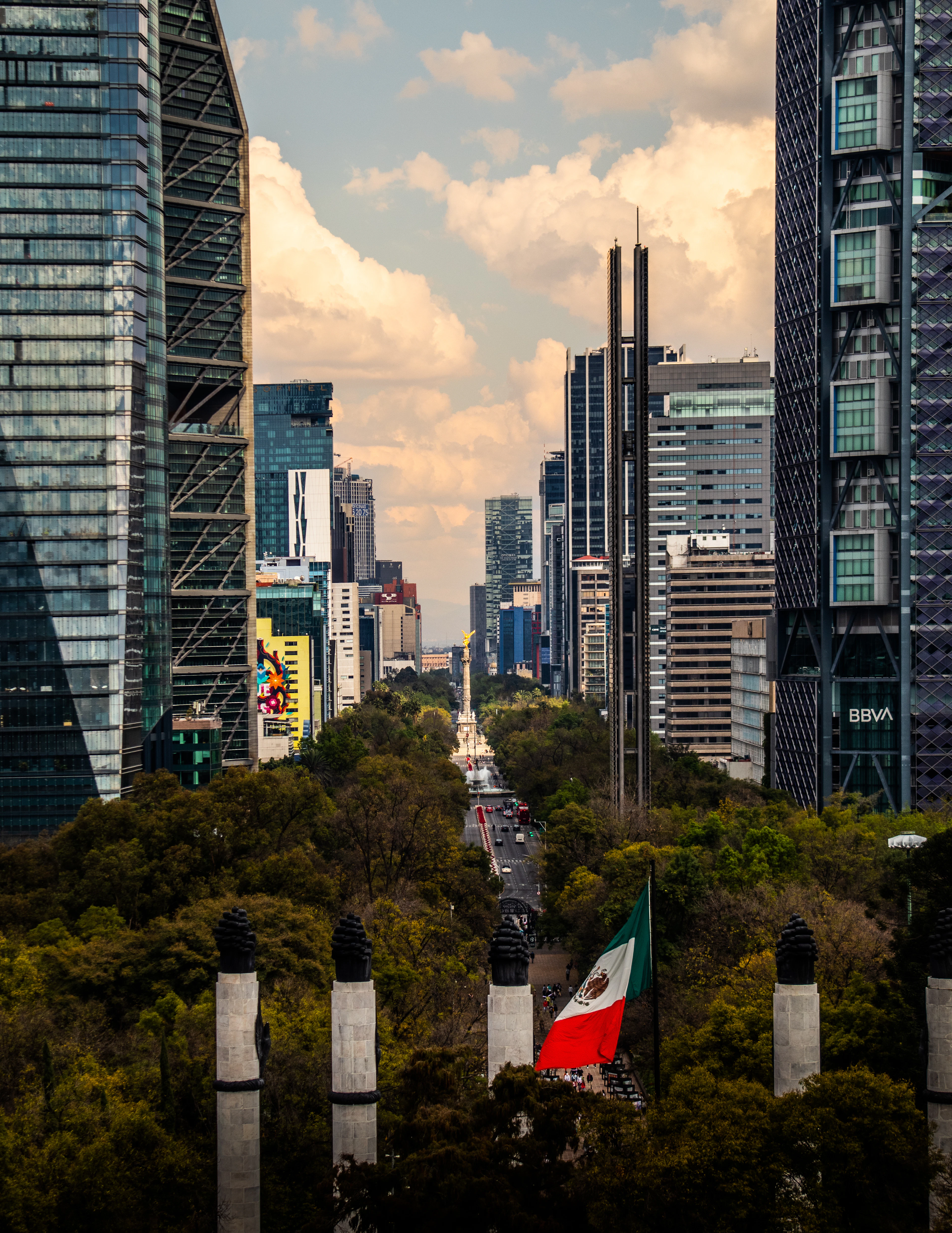 Golden-hour aerial view down Paseo de la Reforma, skyscrapers lining the boulevard with a Mexican flag below.
