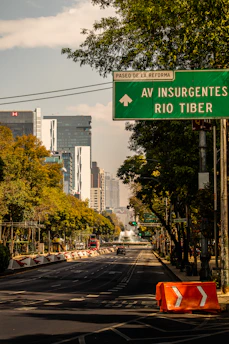 A city street lined with trees leads towards a modern skyline featuring tall buildings. A green directional road sign above the street points towards 'Paseo de la Reforma' and 'Av Insurgentes Rio Tiber'. The road is clear with few vehicles, and there is an orange traffic barrier on the side.
