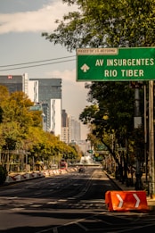 A city street lined with trees leads towards a modern skyline featuring tall buildings. A green directional road sign above the street points towards 'Paseo de la Reforma' and 'Av Insurgentes Rio Tiber'. The road is clear with few vehicles, and there is an orange traffic barrier on the side.