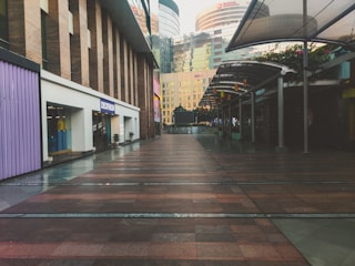 An empty urban shopping complex walkway with modern architectural buildings. The left side features a Decathlon storefront, and the right side has covered walkways with plants on top. The buildings in the background have reflective glass windows.
