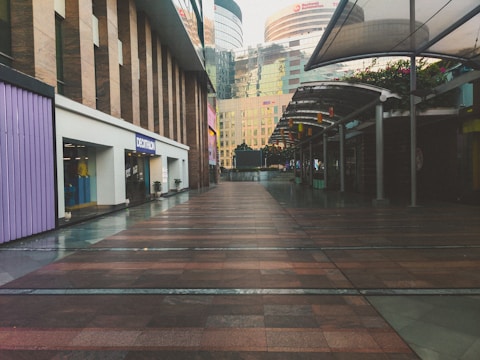 An empty urban shopping complex walkway with modern architectural buildings. The left side features a Decathlon storefront, and the right side has covered walkways with plants on top. The buildings in the background have reflective glass windows.