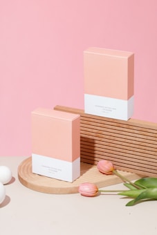 Two peach and white cosmetic boxes are displayed on a wooden platform with a pink background. Tulip flowers and white spheres are placed decoratively around the boxes.