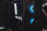 A sleek gaming computer case with visible internal components. The case features RGB lighting with a prominent blue hue emanating from the fans. The mesh panel in front allows a glimpse of the glowing interior, highlighting the modern hardware inside.