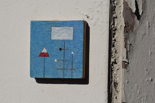 A small square artwork is mounted on a textured, peeling wall. The artwork features a minimalist abstract design with a blue background. It contains geometric shapes including a white rectangle, a red and white triangle, and black lines intersecting with smaller white dots.