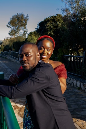 A couple poses outdoors with greenery and a paved pathway in the background. The man wears a dark suit while the woman is dressed in a vibrant red top and headband, smiling warmly.