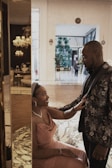 A couple is interacting inside an elegant, well-decorated interior space with marble flooring and warm lighting. The woman, seated and wearing a pink dress, looks up and smiles at the man, who stands beside her in a floral-patterned jacket. The background shows modern decor with chandeliers and a spacious hallway.