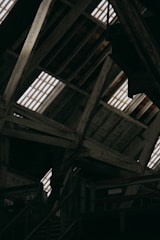 The image displays an interior view of a rustic wooden attic, featuring exposed beams and planks. Light streams through the gaps in the roof, illuminating the weathered wood. The structure appears old and possibly industrial, with metal railings and stairs visible in the lower part of the image.