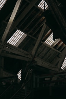 The image displays an interior view of a rustic wooden attic, featuring exposed beams and planks. Light streams through the gaps in the roof, illuminating the weathered wood. The structure appears old and possibly industrial, with metal railings and stairs visible in the lower part of the image.