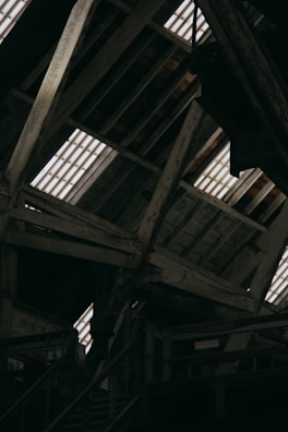 The image displays an interior view of a rustic wooden attic, featuring exposed beams and planks. Light streams through the gaps in the roof, illuminating the weathered wood. The structure appears old and possibly industrial, with metal railings and stairs visible in the lower part of the image.