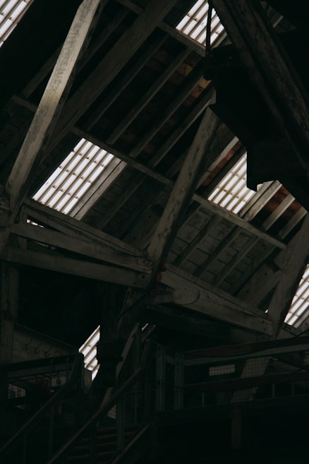 The image displays an interior view of a rustic wooden attic, featuring exposed beams and planks. Light streams through the gaps in the roof, illuminating the weathered wood. The structure appears old and possibly industrial, with metal railings and stairs visible in the lower part of the image.