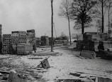 A snowy yard with tall stacks of lumber and wooden planks. Two people are visible, one near a cart filled with wood. The area is surrounded by tall, bare trees indicating a winter setting.