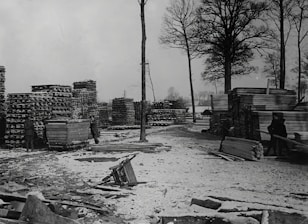 A snowy yard with tall stacks of lumber and wooden planks. Two people are visible, one near a cart filled with wood. The area is surrounded by tall, bare trees indicating a winter setting.