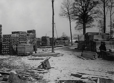 A snowy yard with tall stacks of lumber and wooden planks. Two people are visible, one near a cart filled with wood. The area is surrounded by tall, bare trees indicating a winter setting.