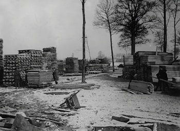 A snowy yard with tall stacks of lumber and wooden planks. Two people are visible, one near a cart filled with wood. The area is surrounded by tall, bare trees indicating a winter setting.