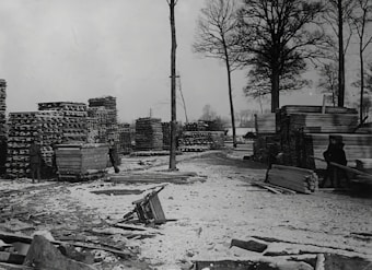 A snowy yard with tall stacks of lumber and wooden planks. Two people are visible, one near a cart filled with wood. The area is surrounded by tall, bare trees indicating a winter setting.