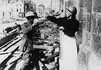 A man and a woman, both wearing vintage clothing and helmets, are standing on a street filled with rubble. The man is holding a large birdcage, while the woman adjusts something on it. The background shows dilapidated buildings, suggesting a scene from a war-torn area.