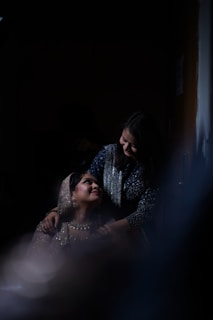Two women are the focus in the image, one sitting and looking up lovingly at the standing woman who has her hands on the seated woman's shoulders. They are dressed in elegant attire, with the seated woman wearing a sparkling outfit that includes a beaded veil over her head. The lighting is dim and dramatic, creating an intimate and warm atmosphere.