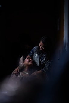 Two women are the focus in the image, one sitting and looking up lovingly at the standing woman who has her hands on the seated woman's shoulders. They are dressed in elegant attire, with the seated woman wearing a sparkling outfit that includes a beaded veil over her head. The lighting is dim and dramatic, creating an intimate and warm atmosphere.