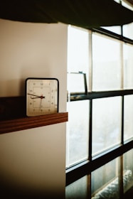 A vintage-style alarm clock sits on a wooden shelf next to a large window. Soft natural light is filtering through the window, creating a calm and serene atmosphere. The clock shows the time as 7:08.