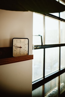 A vintage-style alarm clock sits on a wooden shelf next to a large window. Soft natural light is filtering through the window, creating a calm and serene atmosphere. The clock shows the time as 7:08.