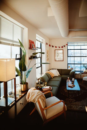 A warm, inviting living room with soft sage green accents, rustic wooden shelves filled with plants and décor, and a cozy knit throw on a cream sofa.