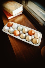 A tray containing a mix of brown and white eggs placed on a wooden surface near a window. Two books with ornate covers are stacked beside the tray, and natural sunlight streams through the window, casting warm shadows.