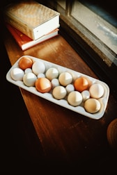 A tray containing a mix of brown and white eggs placed on a wooden surface near a window. Two books with ornate covers are stacked beside the tray, and natural sunlight streams through the window, casting warm shadows.