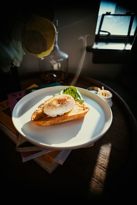 An artistic photograph of a plate containing a slice of toasted bread topped with a poached egg and a side of greens. The setting is dimly lit with sunlight streaming in through a small window, creating a cozy and warm atmosphere. The plate sits on a wooden surface, and there are faint hints of other objects like a glass jar and a stack of books nearby.