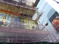 A partially constructed or renovated building covered in bamboo scaffolding with various large printed advertisements and signage visible through and around the scaffolding. Glass buildings surround the site reflecting light. Some signs are written in what appears to be Chinese characters.