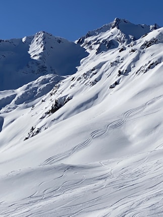 Snow-covered mountains with clear tracks from skiers present, indicating recent activity. The peaks are rugged and dramatic, with a deep blue sky above, enhancing the crispness of the environment.