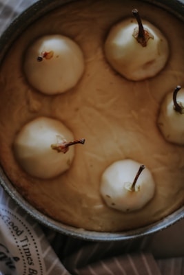 A top view of a baked dessert featuring whole pears with stems intact, arranged upright in a round baking dish. The pears are surrounded by a golden brown pastry or batter. The texture of the baked surface is smooth, with slight browning near the pears.