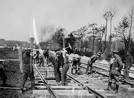 A group of men is working on constructing or repairing railway tracks. Some are using tools like pickaxes, while others appear to be supervising. The setting is outdoors in a rural area, with trees and open land visible in the background. The image is in black and white, indicating it might be from a historical period.