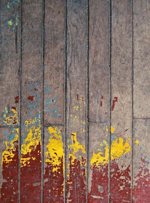 A weathered wooden surface features a pattern of peeling paint in shades of vibrant yellow and red, with traces of blue scattered over the aged brown wood. The paint appears worn, suggesting a rustic or vintage aesthetic.