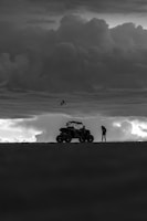 A silhouette of a photographer capturing an off-road vehicle crossing a shallow river, with dark clouds overhead.