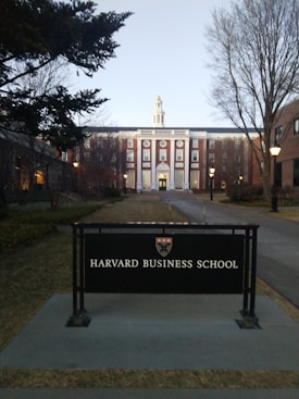 A prominent building with classical architecture featuring a large entrance, evenly spaced windows, and a dome-like structure on top. In the foreground, a sign reads 'Harvard Business School' with a logo above it. Trees and street lamps line the path leading to the building, and it's surrounded by grass.